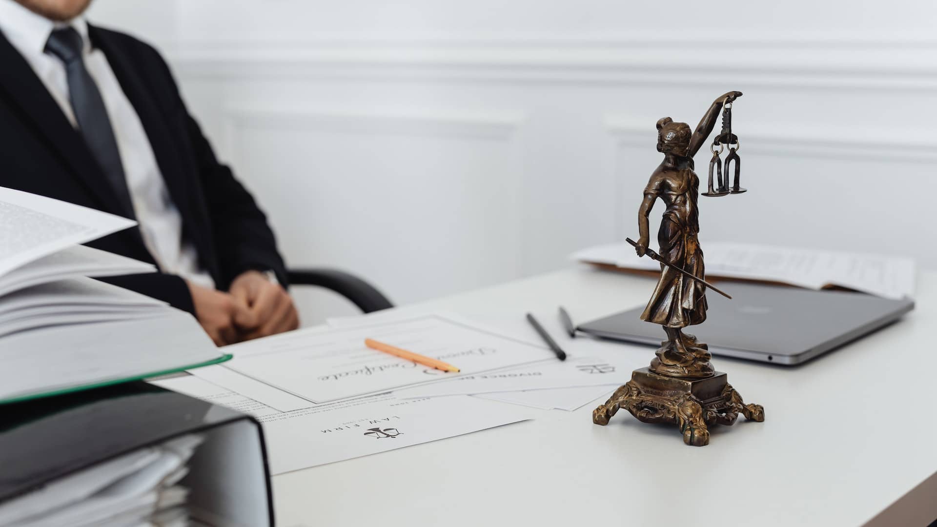 Office desk with a bronze statue of Lady Justice, a laptop, and stationery, with a blurred figure in the background