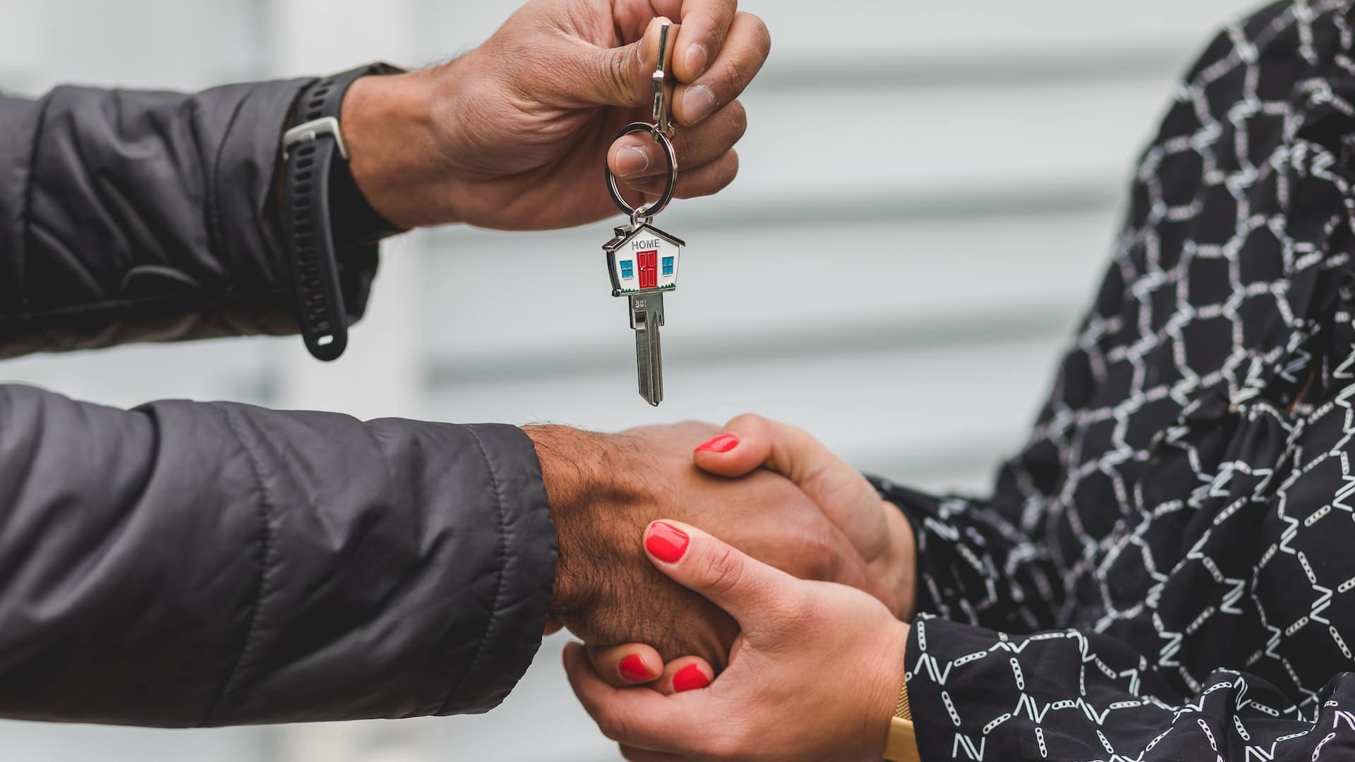 pexels-rdne-stock-project-8293778 Hand in black jacket giving a house key with a small house keychain to a hand in a black and white patterned shirt, against a white wall with horizontal lines